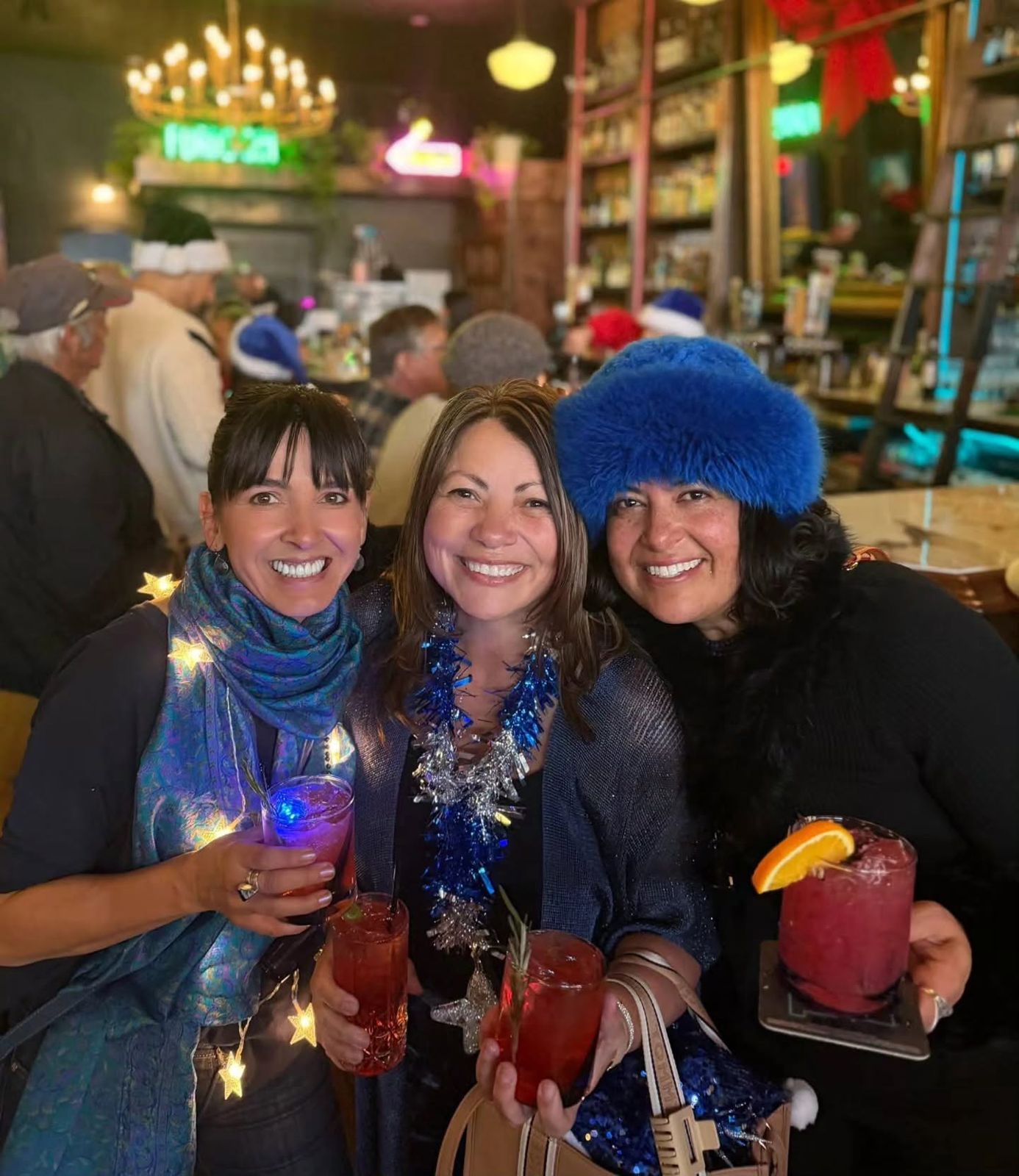 Three friends in festive blue hats and tinsel holding cocktails at Borracho's